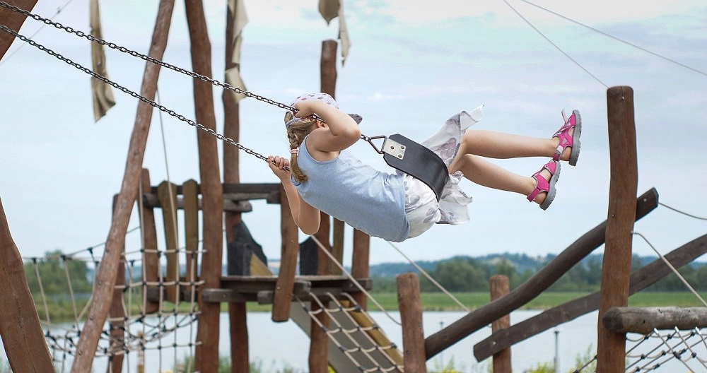 An image showing a child swinging on a belt swing seat with a wooden playset in the background.