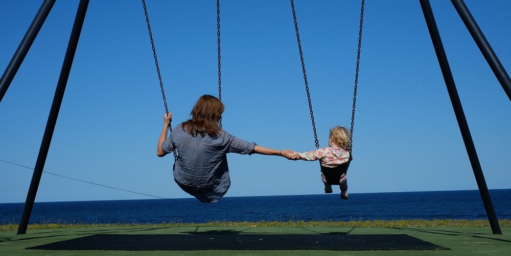 An image showing a woman holding a child's hand while swinging from a swing set.